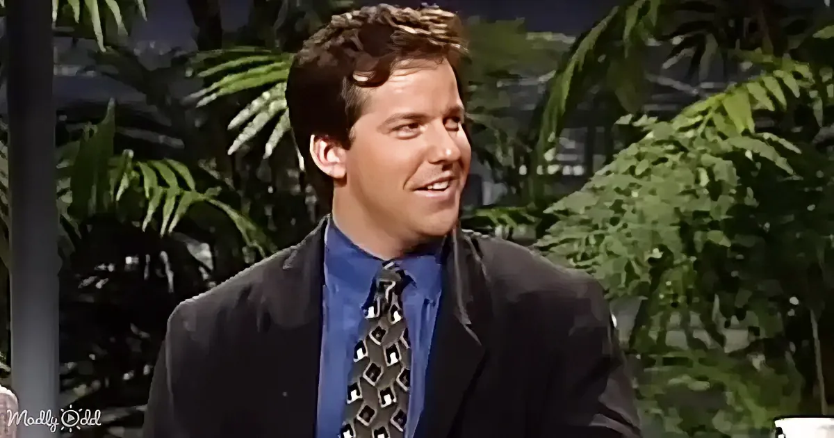 A man in a suit and tie smiles, with tropical plants in the background.