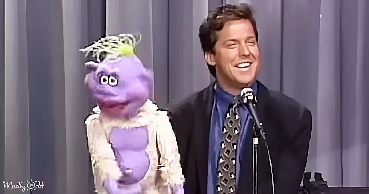 A man in a suit and tie is smiling next to a purple puppet with a white fur collar.