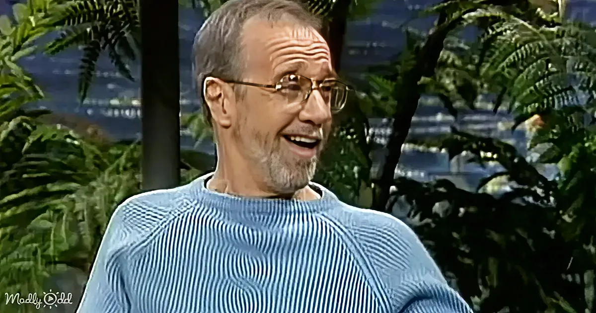 A man in a blue striped sweater smiles while wearing glasses, surrounded by greenery.
