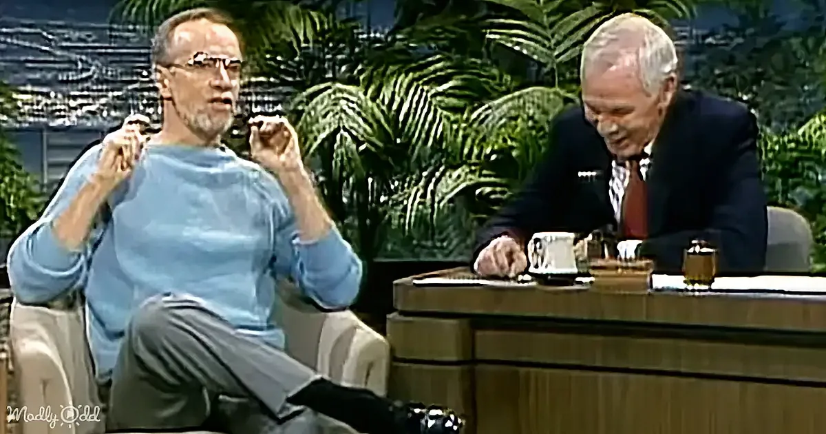A man in a blue sweater gestures animatedly while seated, with another man in a suit observing at a desk.