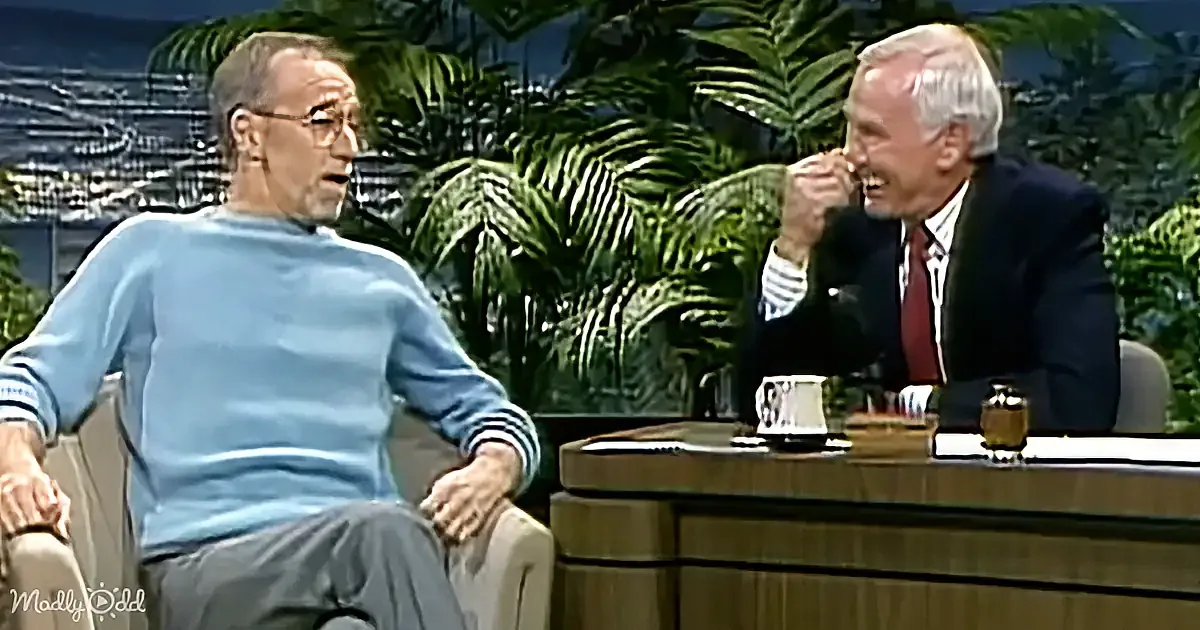 A man in a blue sweater sits on a chair, engaged in conversation with another man in a suit at a desk.