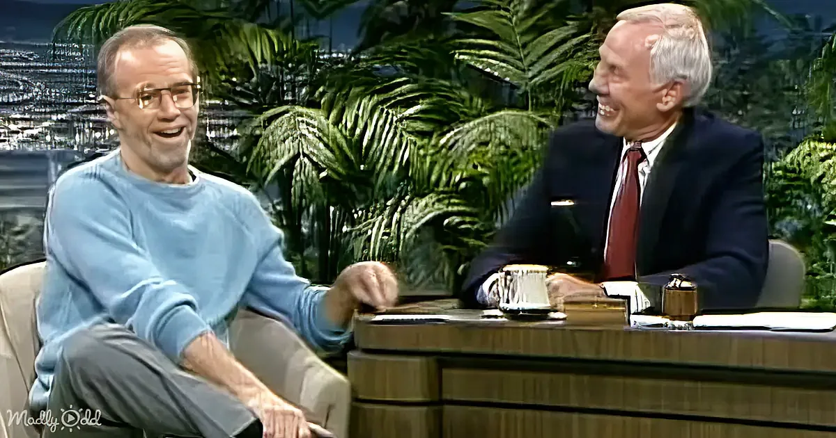 Two men are seated, one in a blue sweater and the other in a suit, both smiling and engaged in conversation.