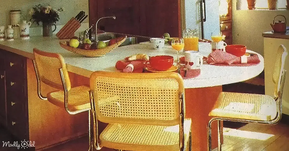 A retro kitchen with wooden cabinets, a white countertop, and woven chairs around a dining table set for breakfast.