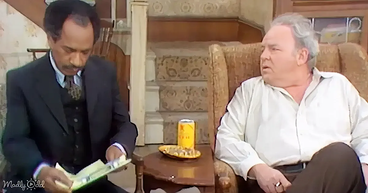 A man in a suit reads a book while another man in a white shirt sits on a couch, both in a cozy room setting.