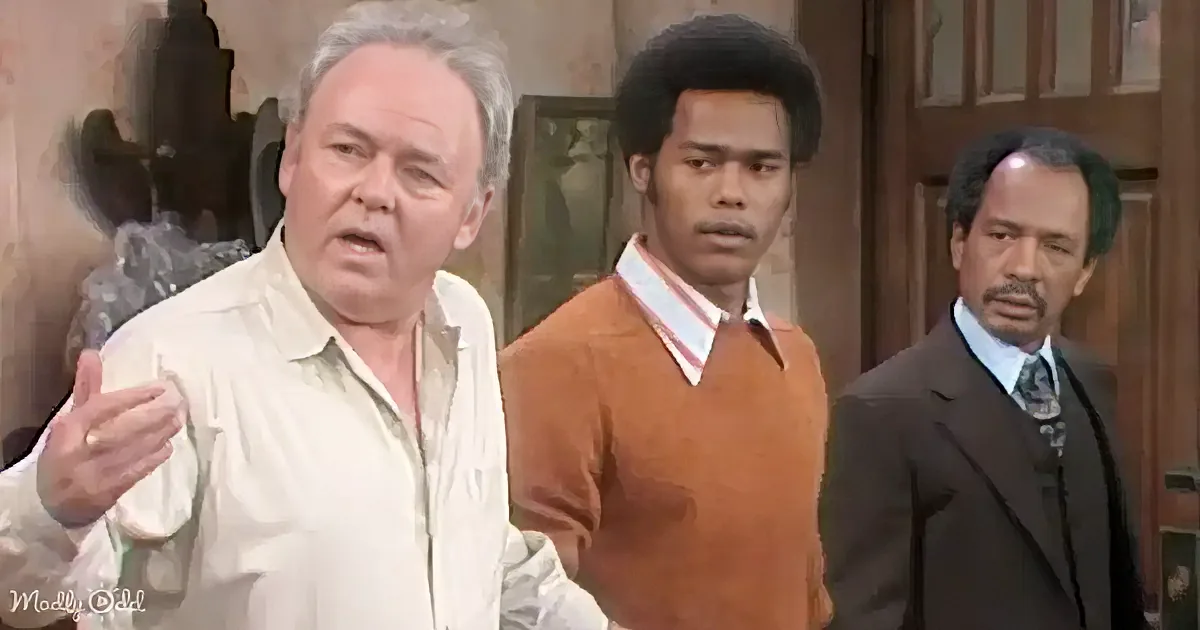 A man in a white shirt gestures while two others, one in a brown sweater and another in a suit, look on.