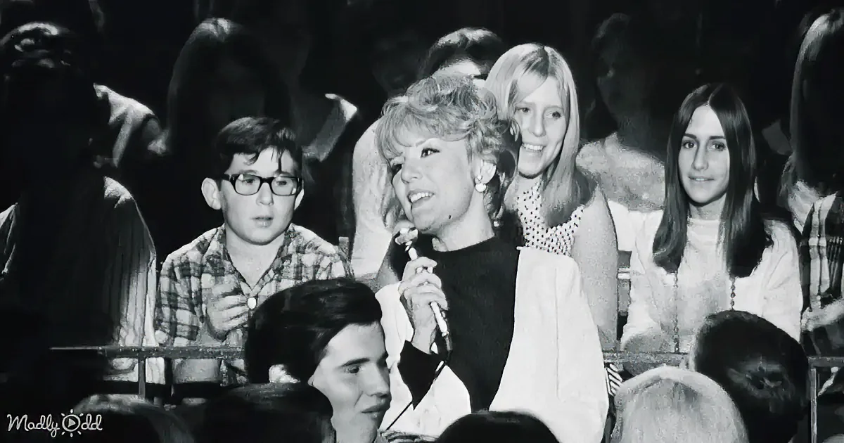 A woman in a white suit holds a microphone, standing in front of a blurred audience.