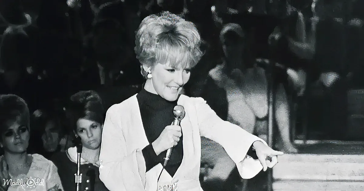 A woman in a white outfit holds a microphone, smiling, with a band and audience in the background.