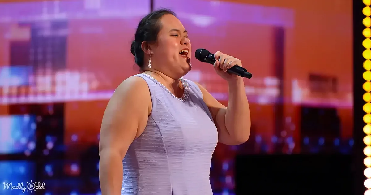 A woman in a white dress sings passionately into a microphone on a stage with a cityscape backdrop.