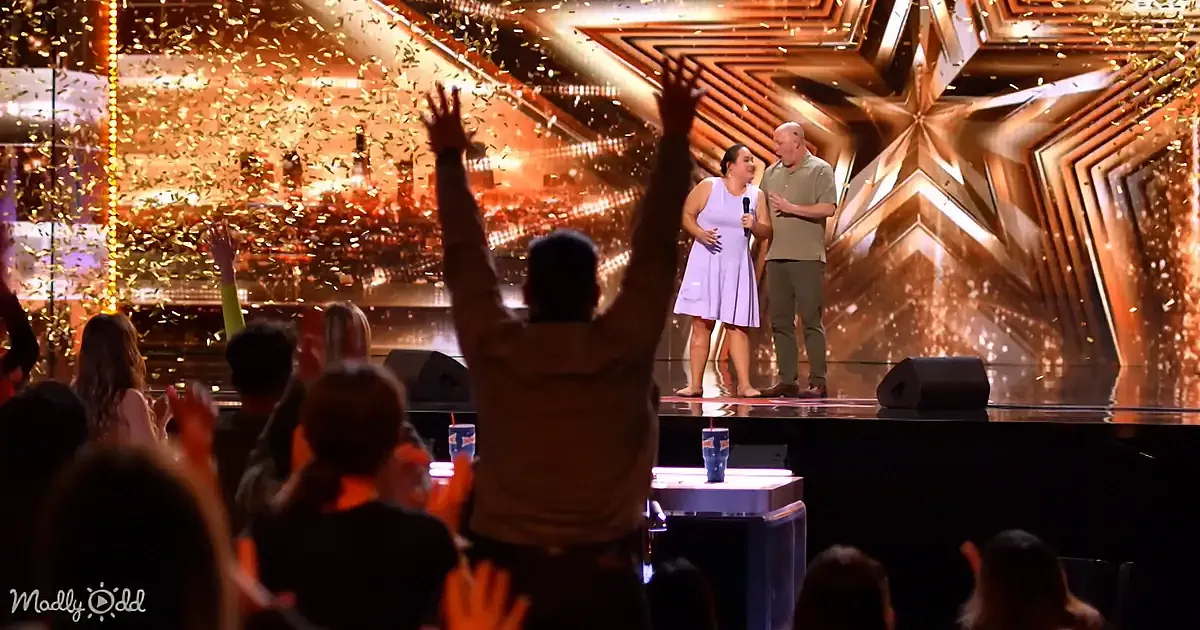 A woman in a white dress stands on stage with a man, surrounded by confetti and a cheering audience.