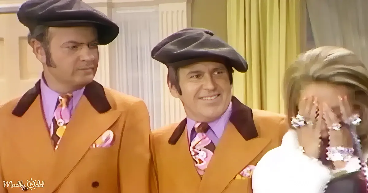 Two men in orange suits and berets stand side by side, smiling, with a woman in the background covering her face.