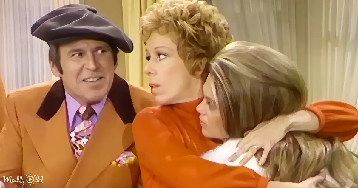 A woman in an orange dress embraces another woman while a man in a beret and orange suit looks on.