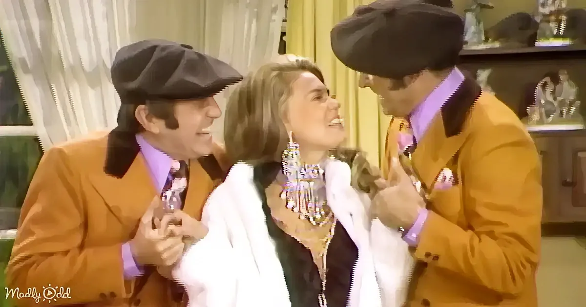A woman in a white coat and jewelry is flanked by two men in matching orange suits and black hats, both smiling.