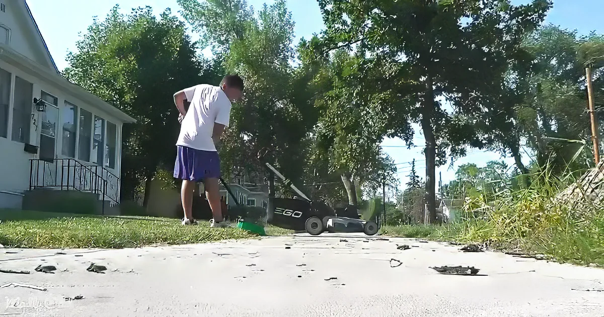 A man mows grass with a black EGO lawnmower on a sunny day near a house.