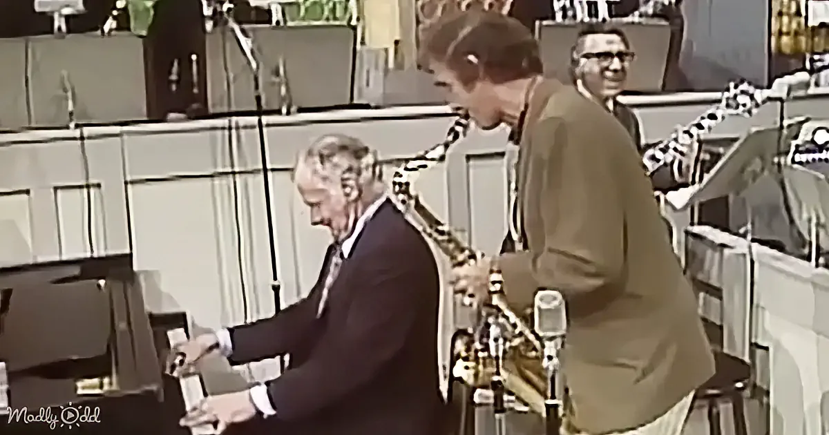A man in a suit plays piano while another man in a brown suit stands nearby, possibly a guest or interviewer.