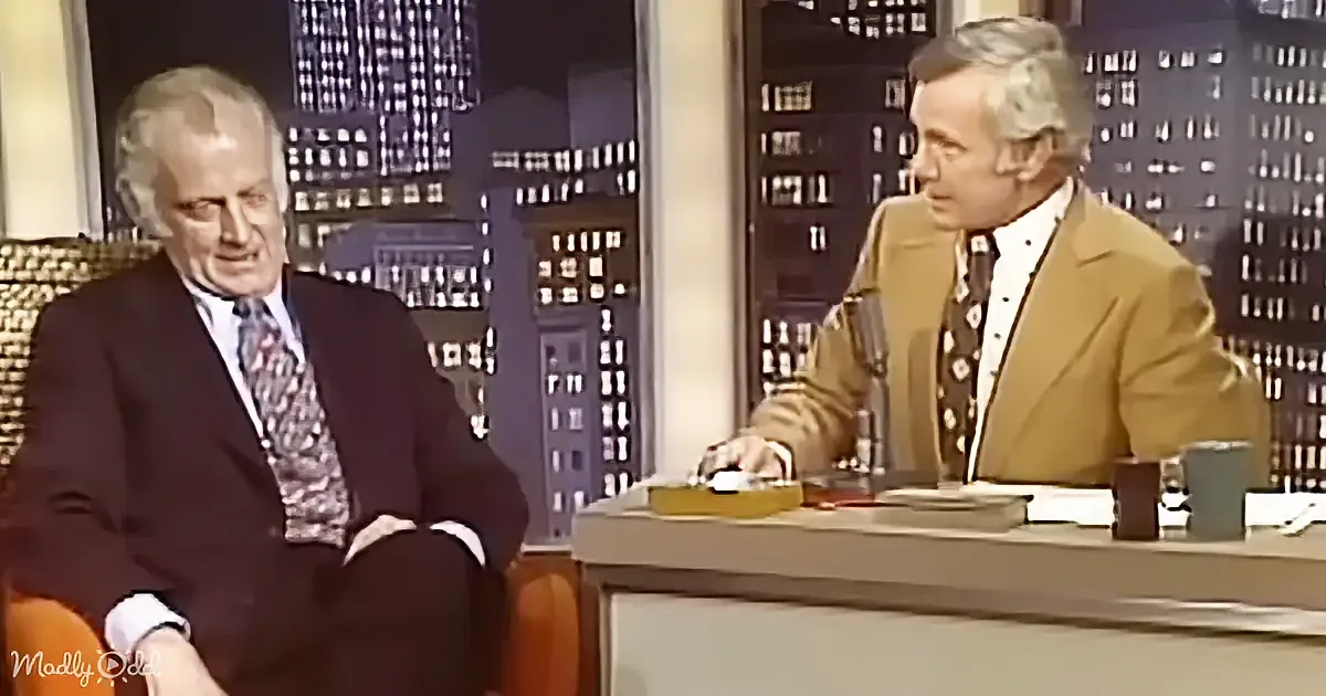 Two men in suits sit on a talk show set with a cityscape backdrop.