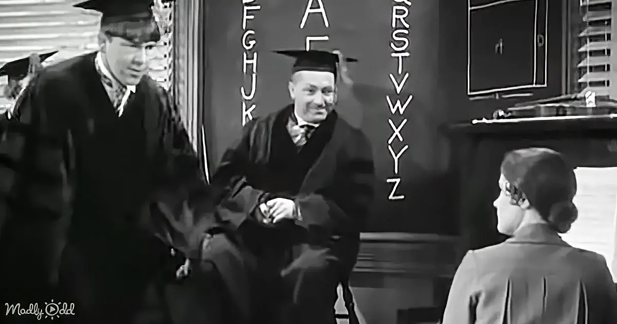 A man in graduation attire sits at a desk with letters on the wall, while another man in a suit stands nearby.