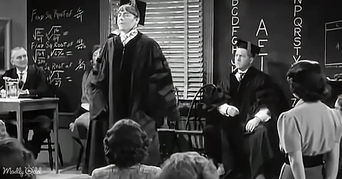 A man in a graduation gown stands at a podium in a classroom, with a chalkboard behind him.
