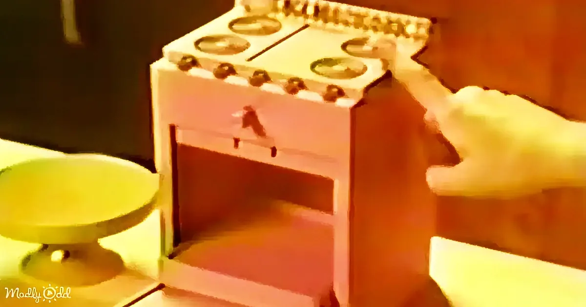 A hand reaches towards a miniature toy oven with a green bowl nearby.