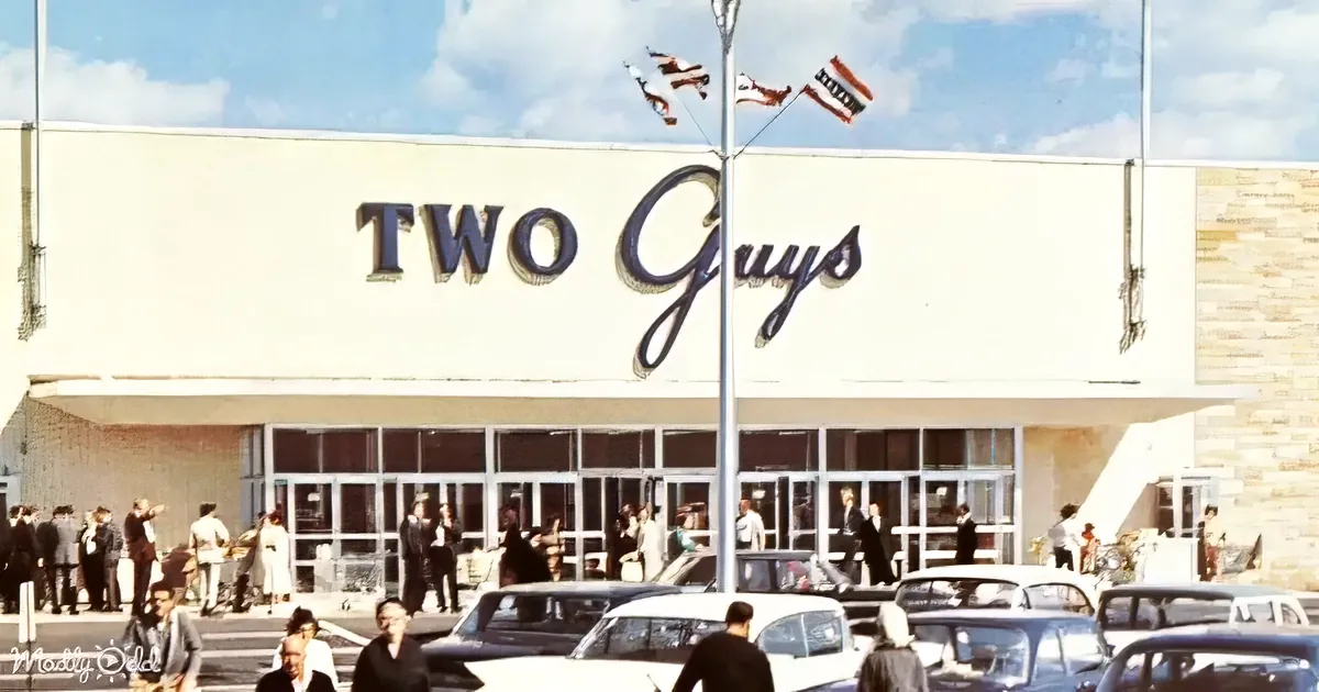 Two Guys store with people outside, vintage cars, flags flying, and a clear sky.