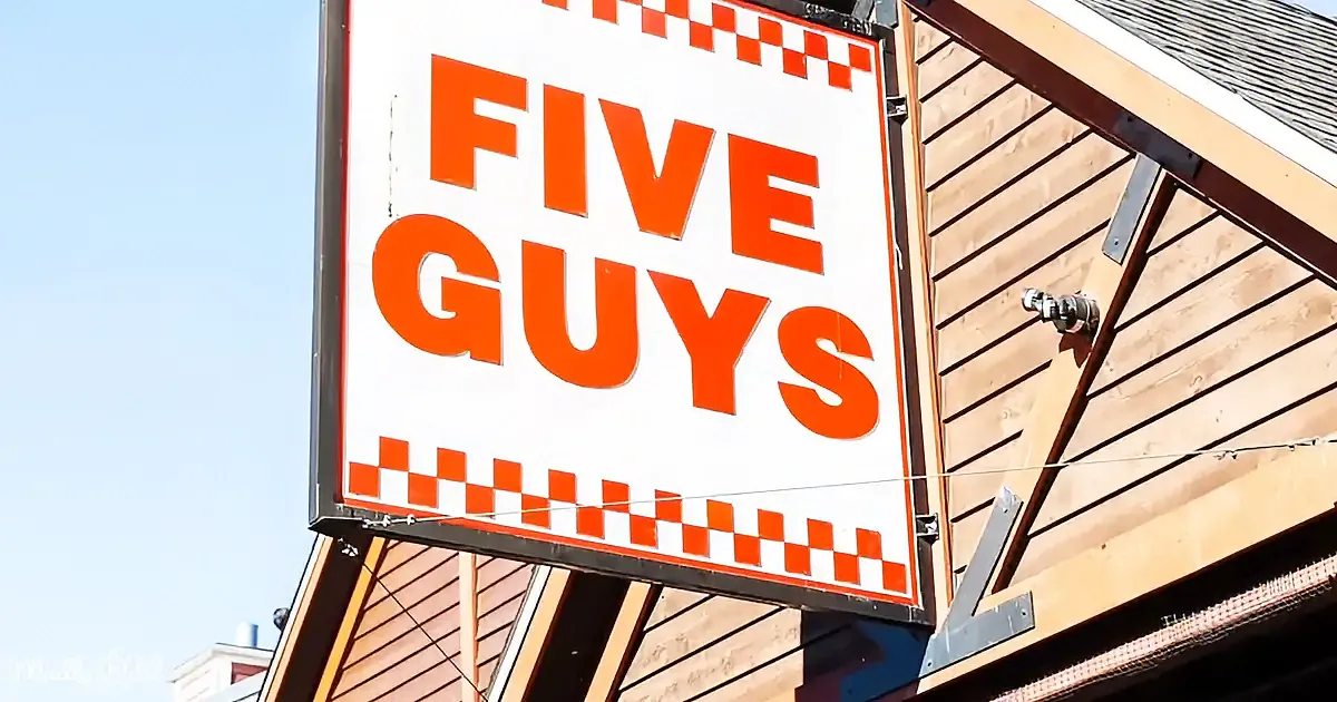 A Five Guys sign with orange text and red checkered border is mounted on a wooden building.
