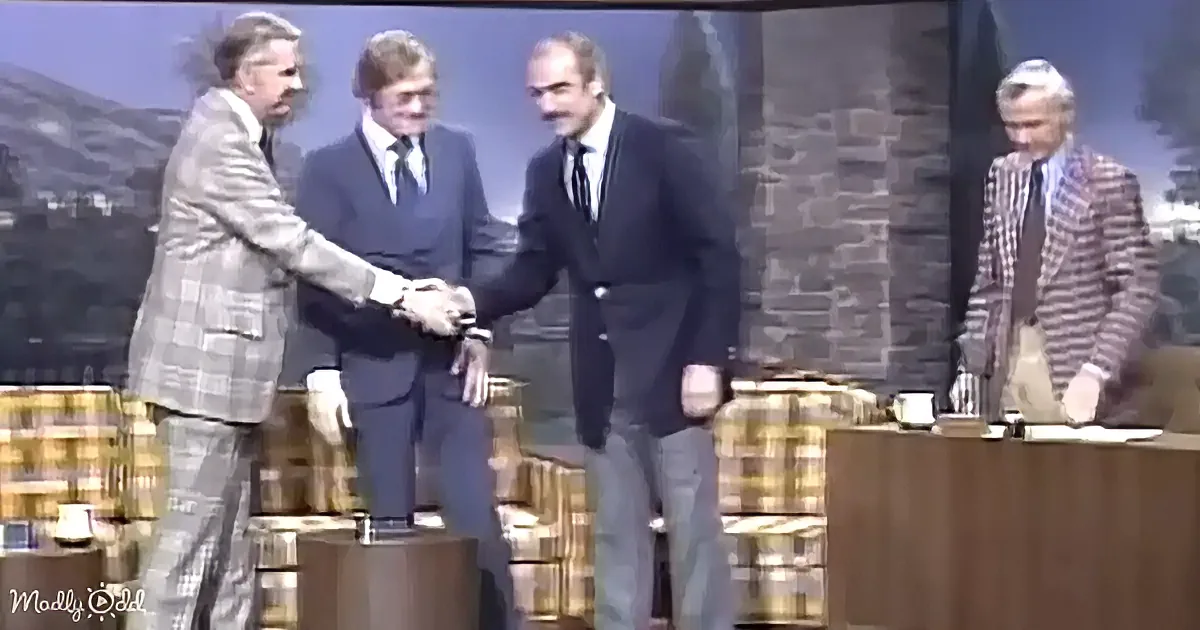 Three men in suits shake hands on stage, one in plaid suit, another in dark suit, and the third in a checkered suit.