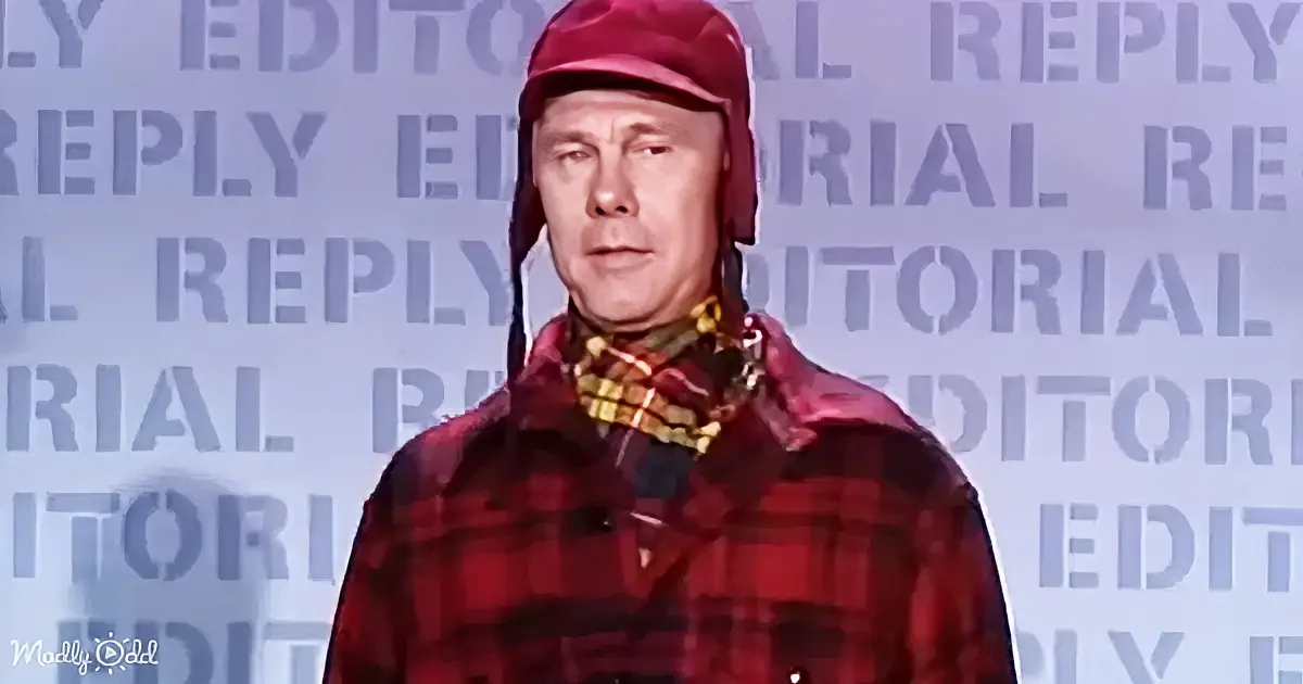A man in a red plaid shirt and aviator hat stands against a backdrop with repeated "EDITORIAL REPLY" text.