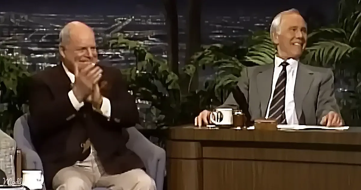 Two men in suits, one clapping, the other smiling, on a talk show set with a cityscape backdrop.