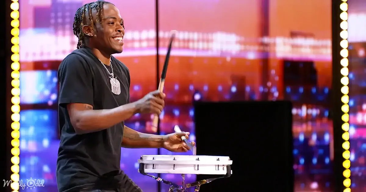 A man in a black shirt smiles while juggling with a white object and a stick on a stage with colorful lights.