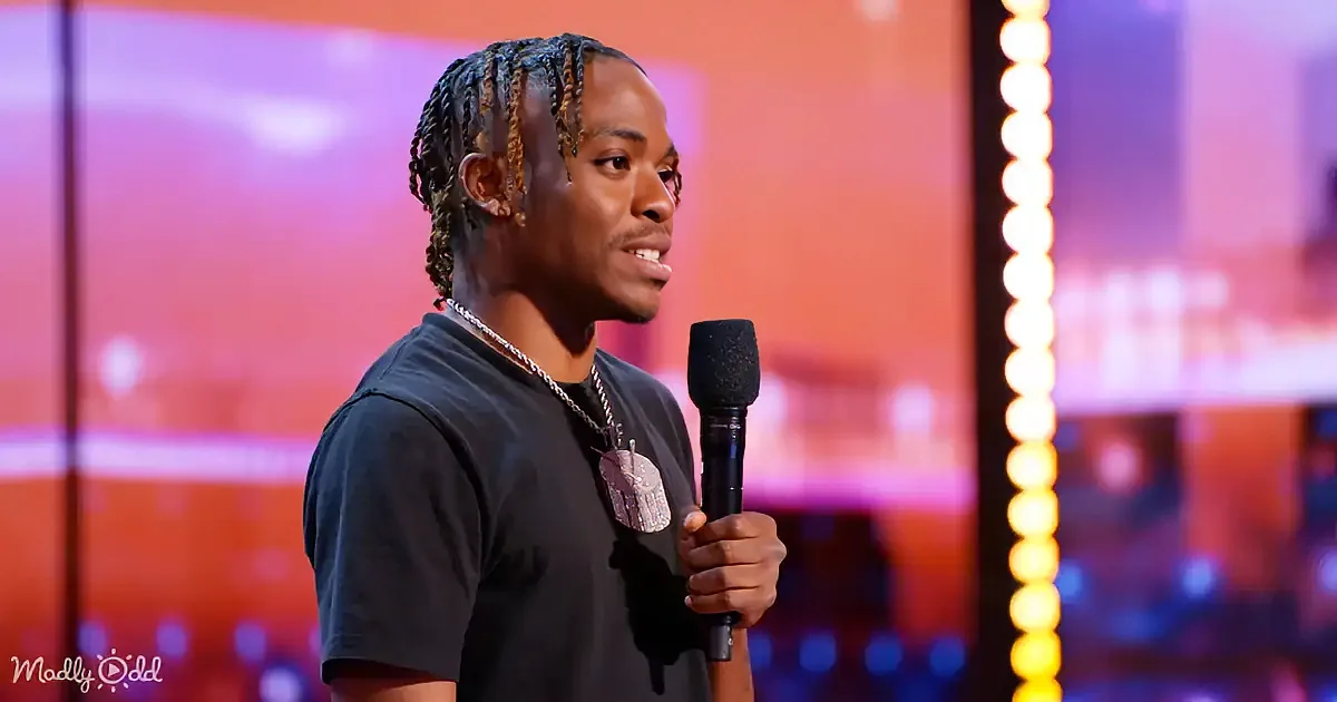 A man with braids holds a microphone, wearing a black shirt and necklace, against a colorful background.