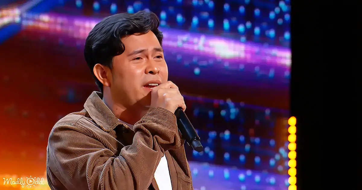 A man in a brown corduroy jacket sings into a microphone on a stage with colorful lights.
