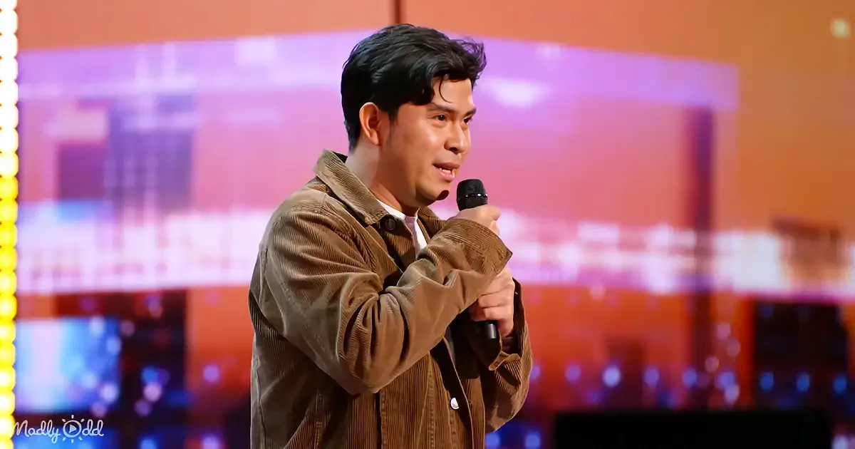 A man in a brown corduroy jacket holds a microphone, speaking on stage with a colorful background.