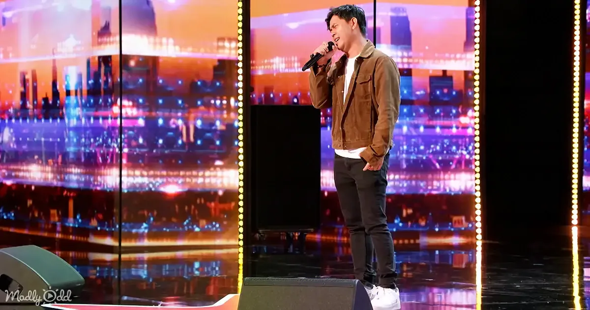 A man in a brown jacket sings into a microphone on a stage with a cityscape backdrop.