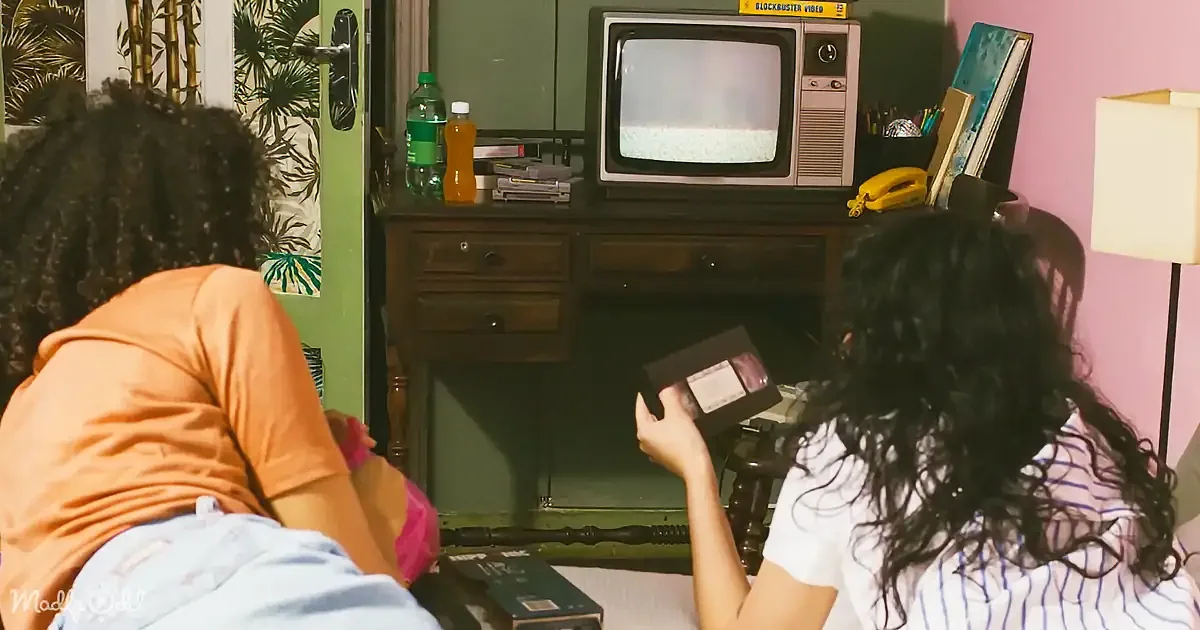 Two individuals are in a room with a vintage TV, a desk, and a lamp, one holding a VHS tape.