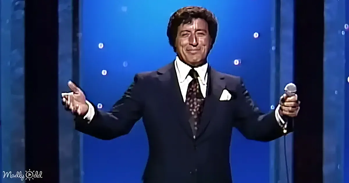 A man in a dark suit and tie holds a microphone and gestures with his other hand on stage.