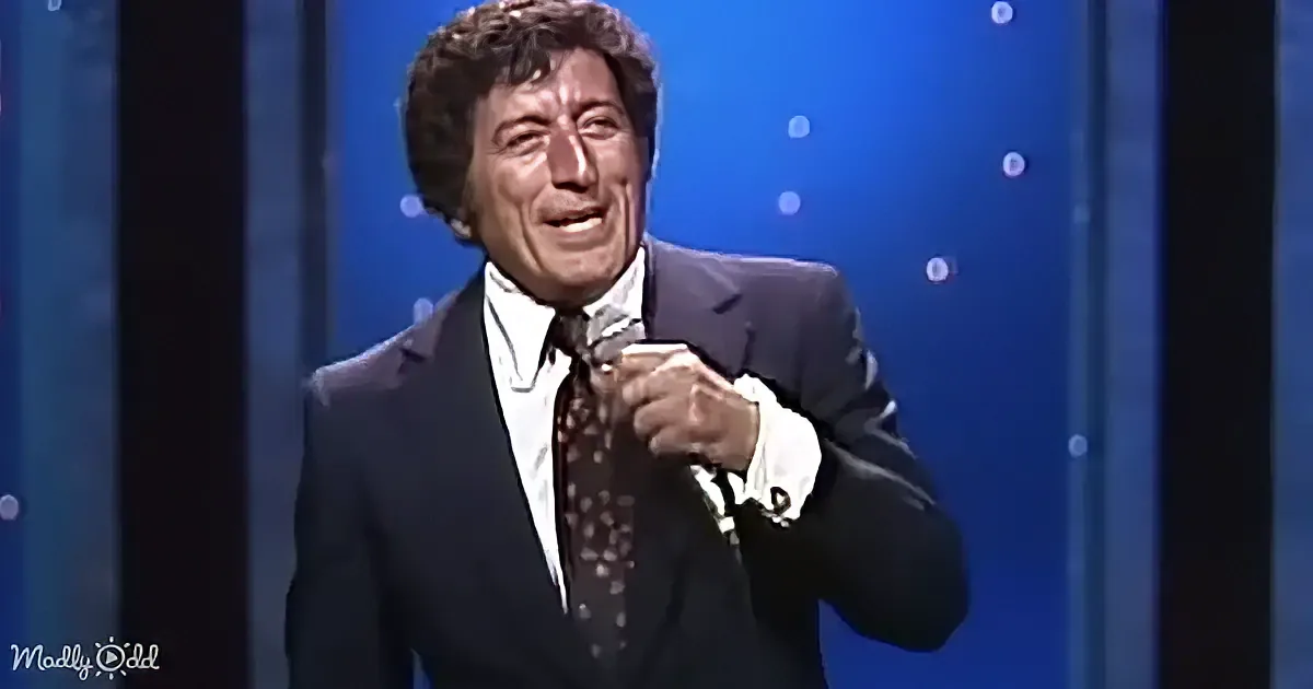 A man in a suit and tie is holding a microphone, smiling, with a blue background.