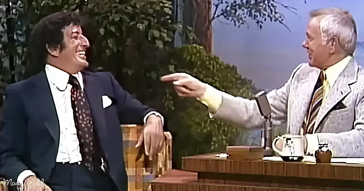 A man in a suit and tie is laughing, pointing at another man in a suit, who is seated behind a desk.