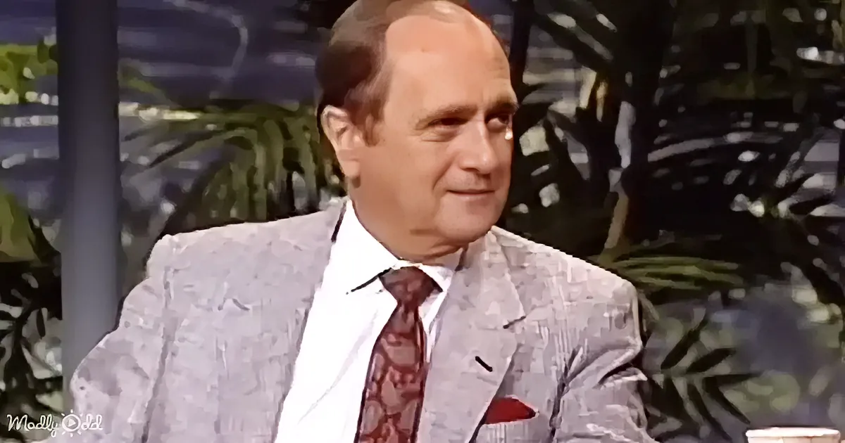 A man in a light gray suit with a red tie and pocket square sits against a backdrop of green foliage.