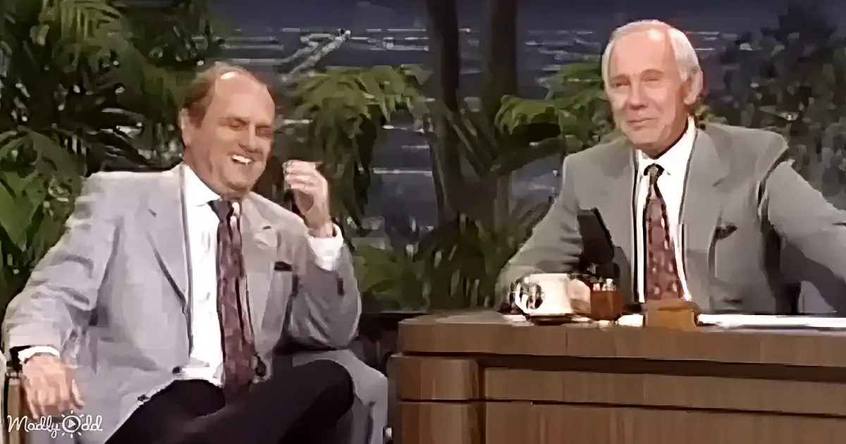 Two men in suits sit on a talk show set, one gesturing animatedly while the other listens.