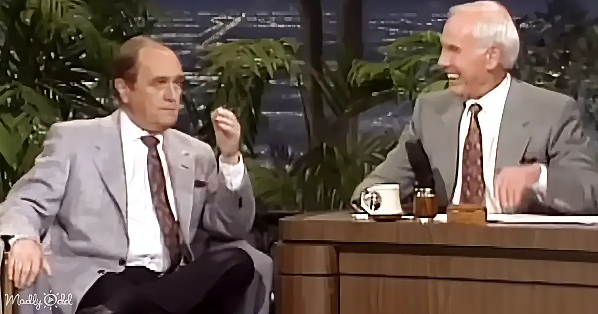 Two men in suits sit at a desk, one gesturing while the other smiles, with a backdrop of plants and city lights.