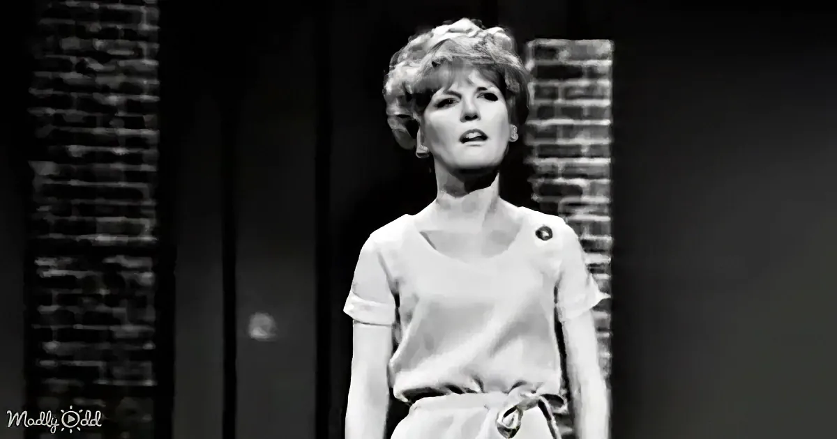 A woman in a white outfit stands between brick walls, looking upwards with a serious expression.