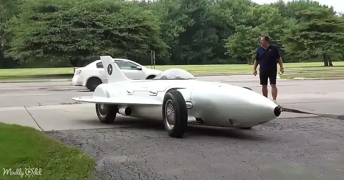 A futuristic white car with a sleek, aerodynamic design is parked on a driveway, with a man standing nearby.