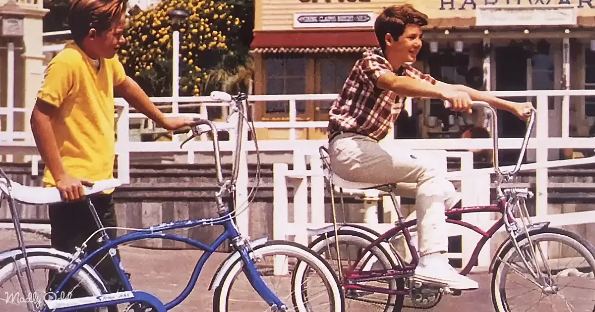 Two boys on bicycles, one in yellow shirt, the other in plaid shirt, in front of a building with a sign.