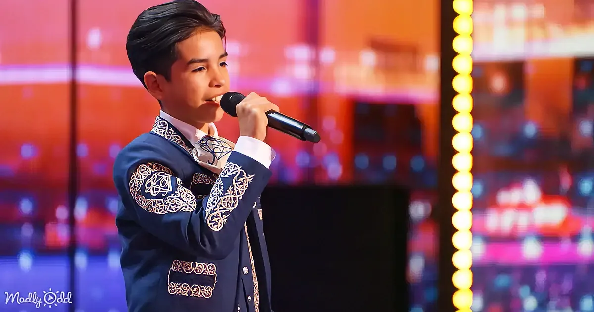 A young boy in a blue embroidered suit sings into a microphone on a stage with colorful lights.
