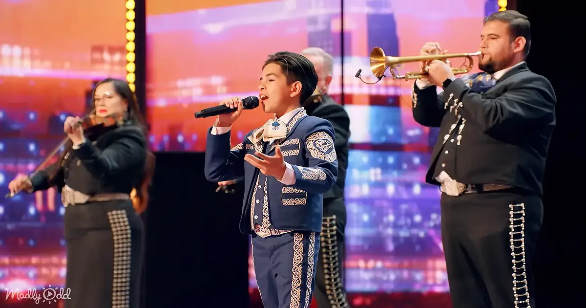 A young boy in a blue mariachi outfit sings into a microphone on stage.