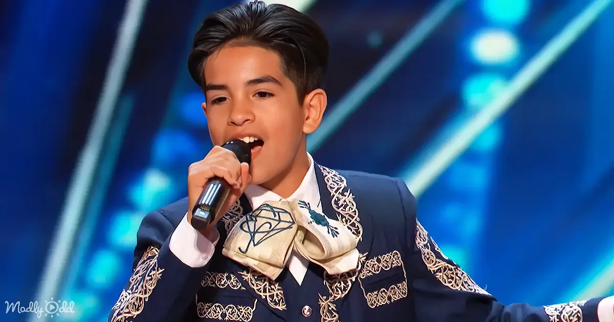 A young person in a traditional embroidered outfit sings into a microphone on a stage with blue lighting.