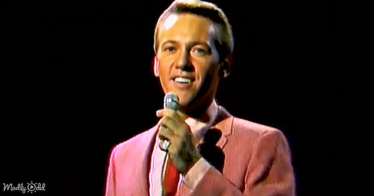 A man in a pink suit holds a microphone, smiling against a dark background.