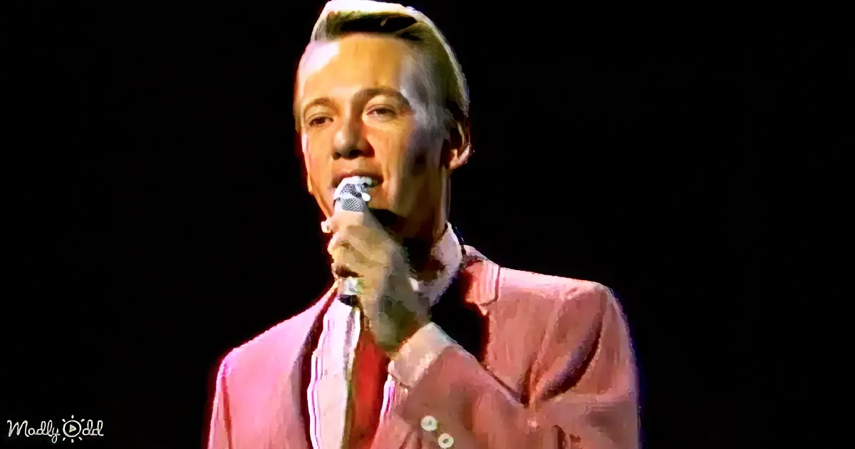 A man in a pink suit sings into a microphone against a dark background.