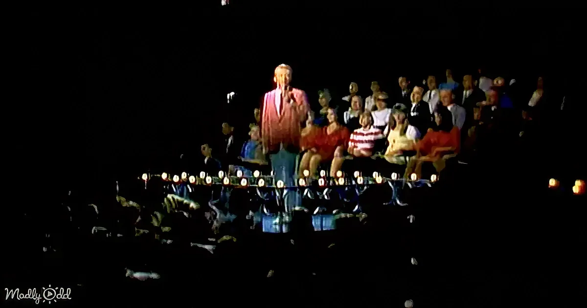 A man in a suit stands on a stage with a microphone, surrounded by seated figures in a dimly lit setting.