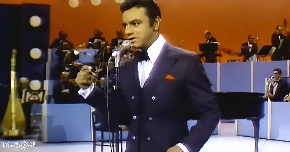 A man in a suit sings into a microphone on stage with an orchestra behind him.