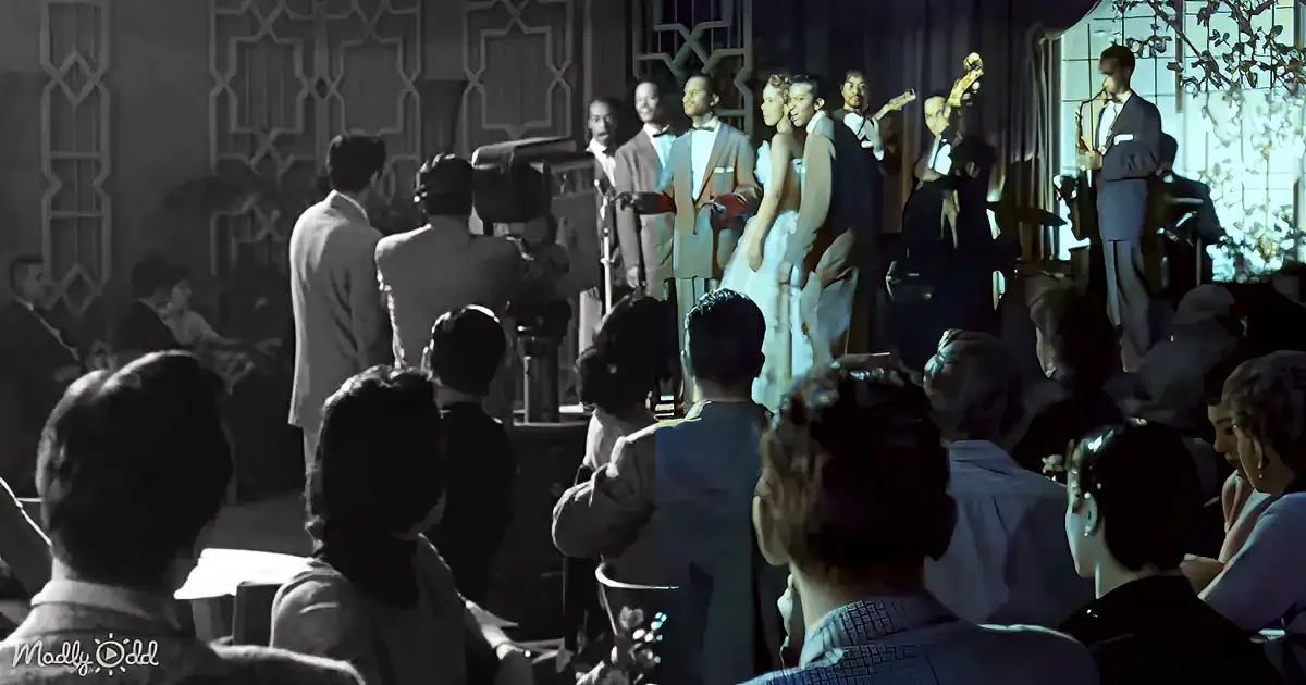 A bride and groom stand on stage, surrounded by musicians and an audience, in a dimly lit, elegant setting.